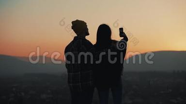恋爱中的年轻夫妇在山上欣赏日落的<strong>剪影</strong>。 他们在智能<strong>手机</strong>上拍照。 假期